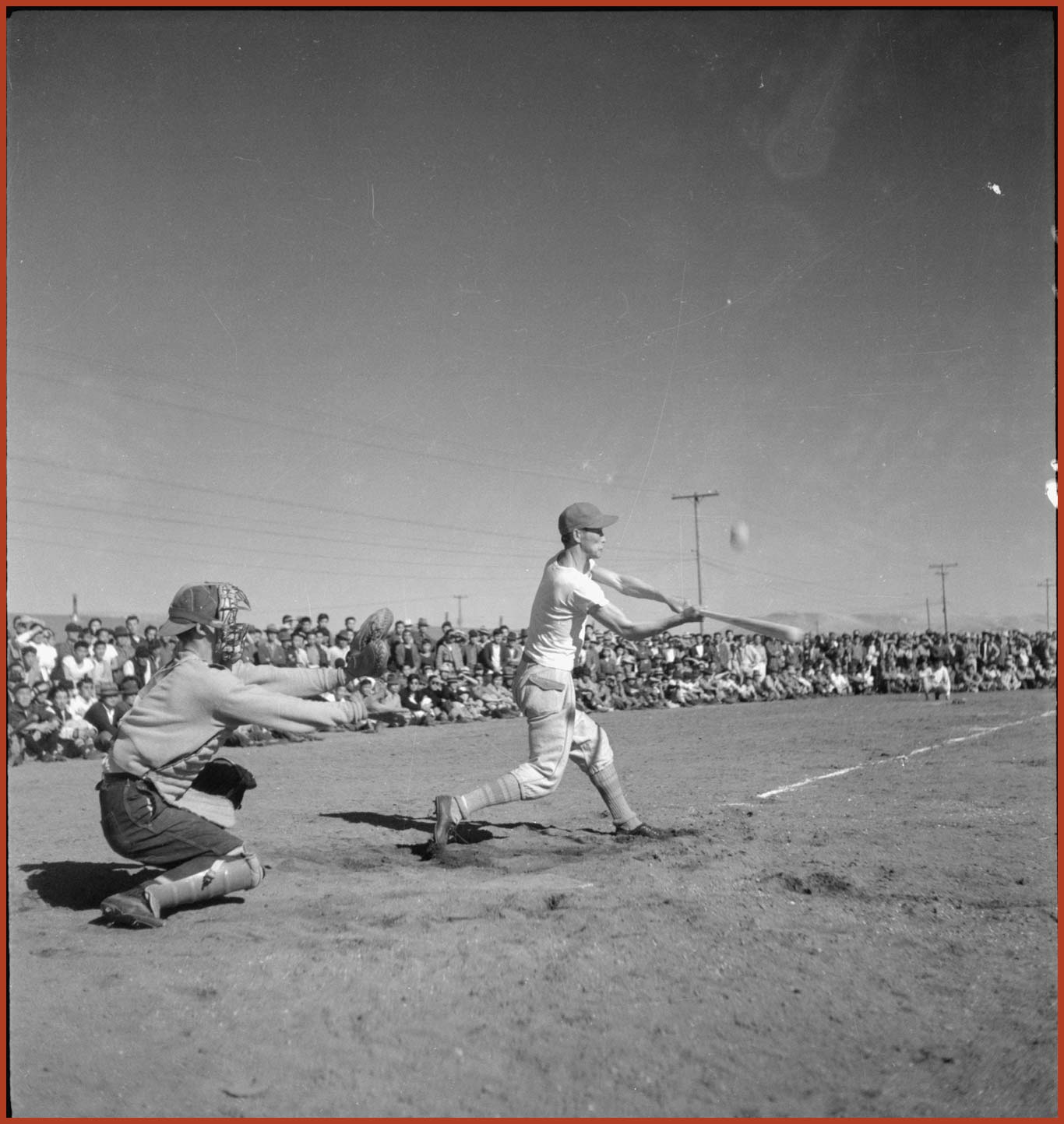 A black and white photograph of two male subjects on a baseball field and an audience in the background. The male two subjects are playing base ball. One male subject is hitting the ball while the other is a catcher behind him.
