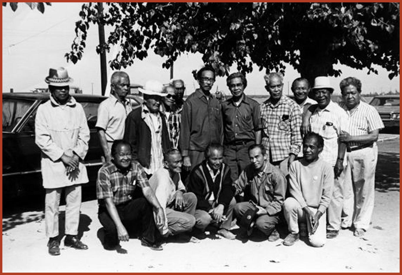 A group of 15 men dressed in farm-work clothes standing in front of 2 cars posing for a photo.