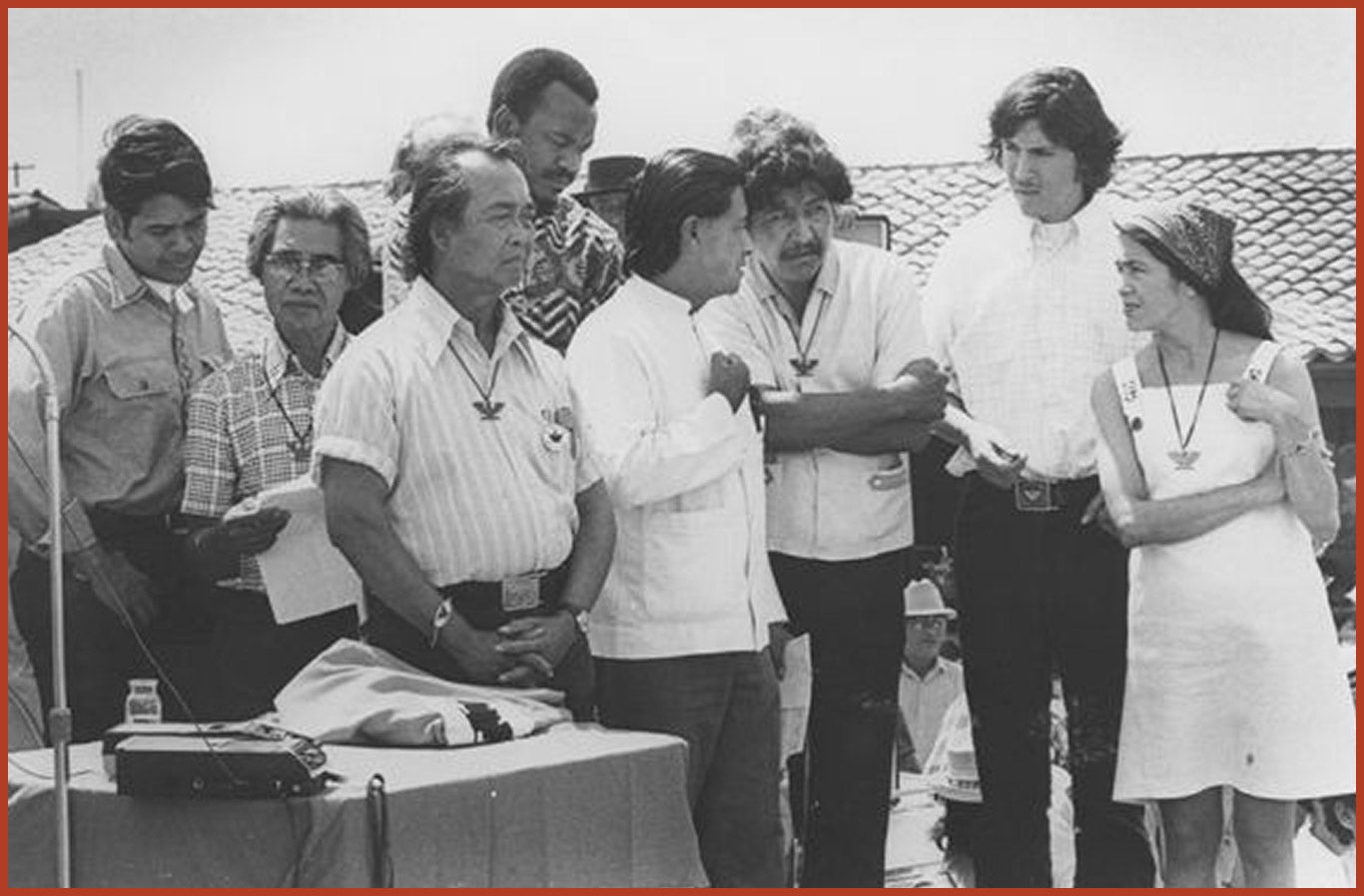 A group of 8 individuals standing outside of a building in conversation. They are all wearing an eagle emblem around their necks.