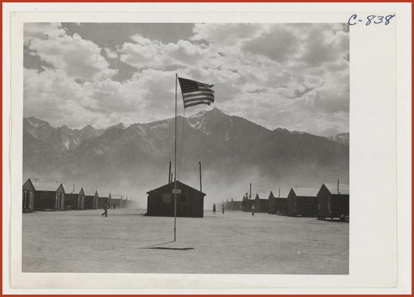 Street scene of barrack homes at this War Relocation Authority Center. The windstorm has subsided and the dust has settled. In the foreground, the American flag waves on a flag pole.