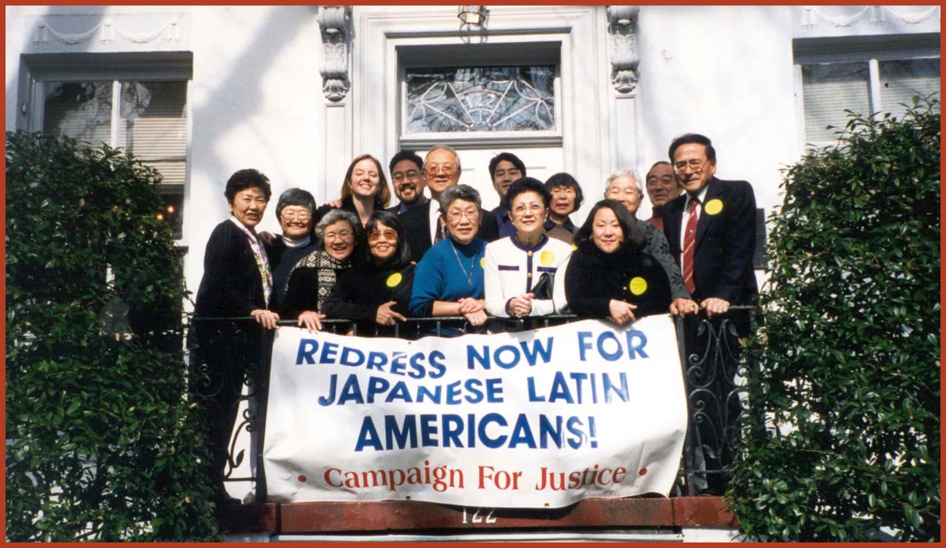 A group of people dressed in business formal attire standing on a balcony holding up a banner with the words "REDRESS NOW FOR JAPANESE LATIN AMERICANS: CAMPAIGN FOR JUSTICE."