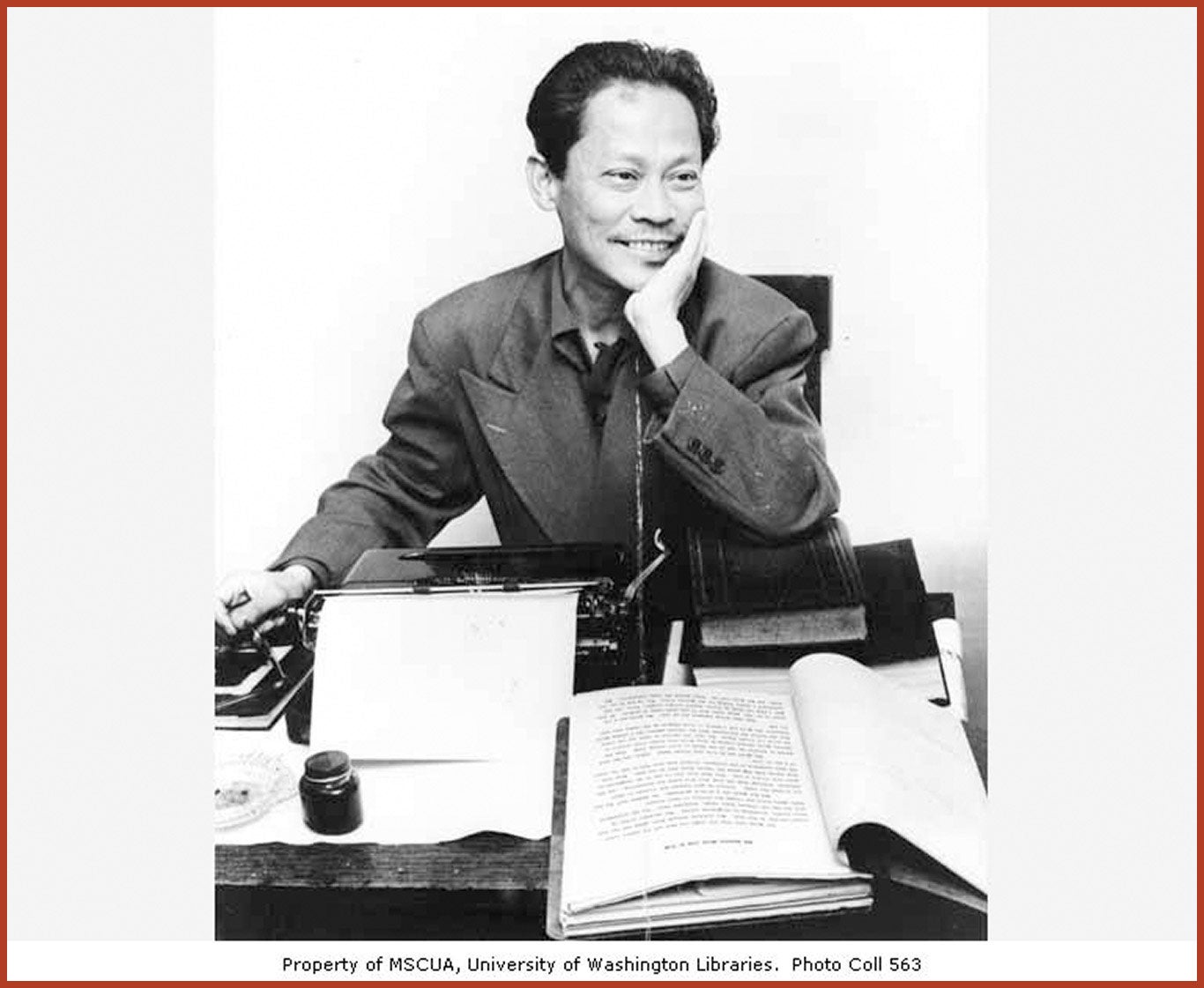Carlos Bulosan, Filipino-American writer, sits at a writing desk with his hand on his cheek, smiling. A typewriter and stacks of books are visible on the desk.