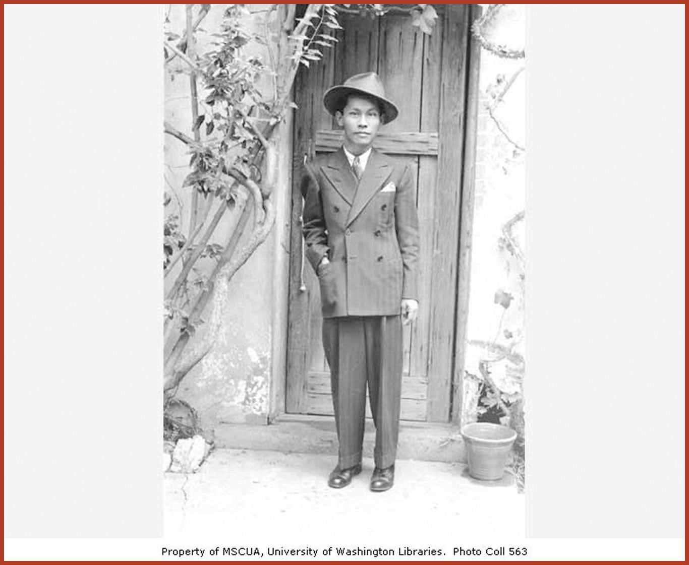 Carlos Bulosan in a suit and hat standing in front of a wooden door.