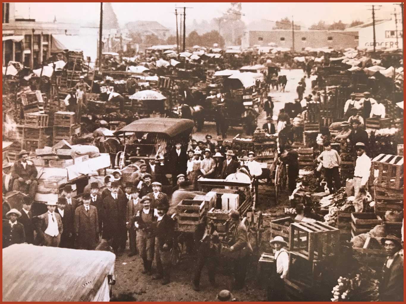 Black and white photo of the Los Angeles City Market. It is crowded with people, cars, wooden crates, and tables.