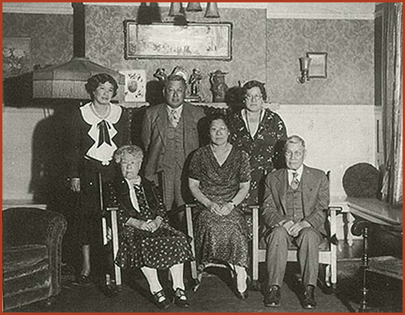 Family portrait of the Tape family in the late 1920s in their living room. Mary, Mamie, and Joseph are sitting while Gertrude, Frank, and Emily are standing behind them. Joseph and Frank are dressed in suits while Mary, Mamie, and Gertrude are wearing long dresses.