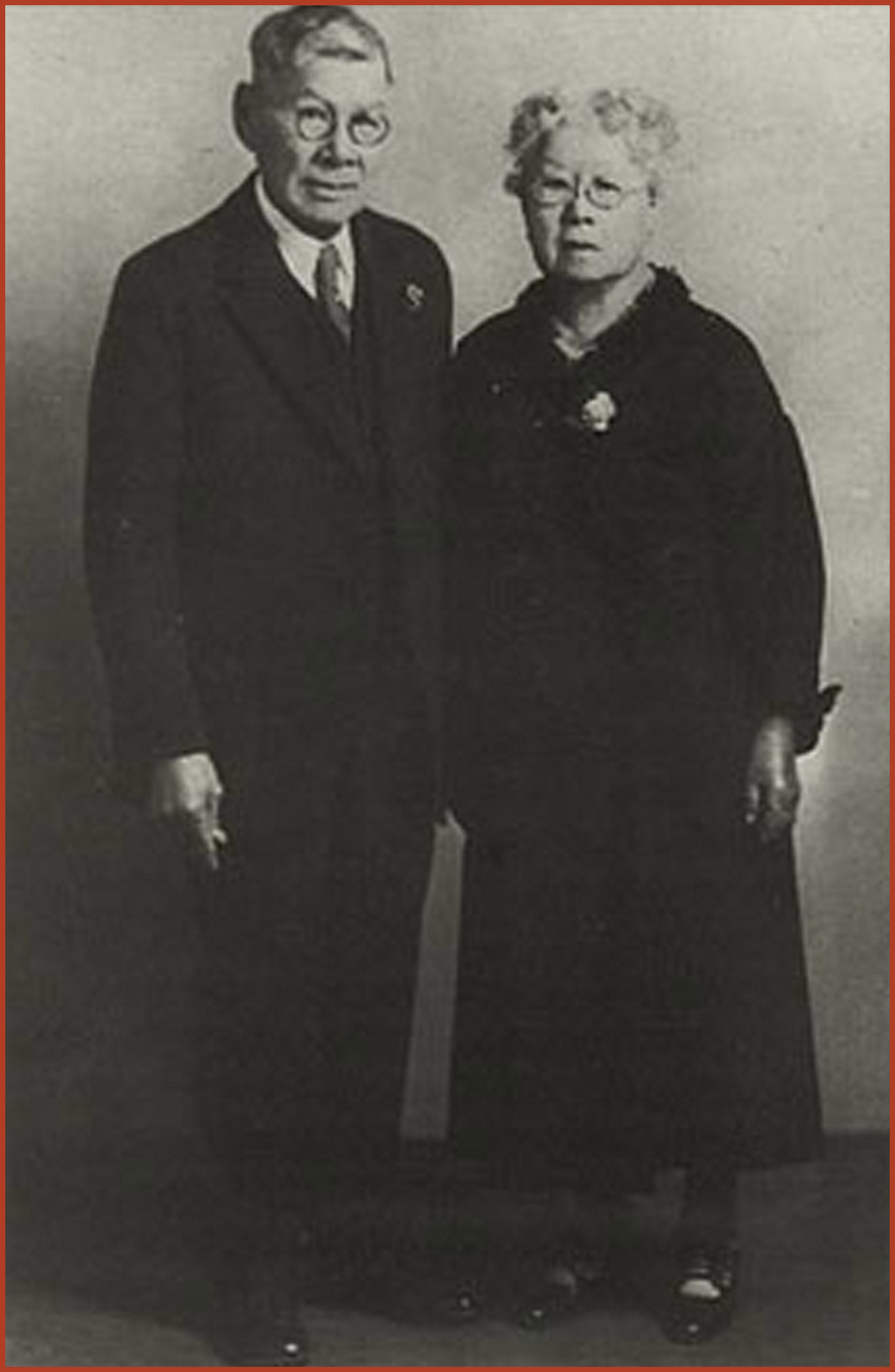 Mary and Joseph Tape, captured in black and white. Joseph Tape is wearing a black suit and circular glasses. Mary Tape is wearing a long black dress and circular glasses.