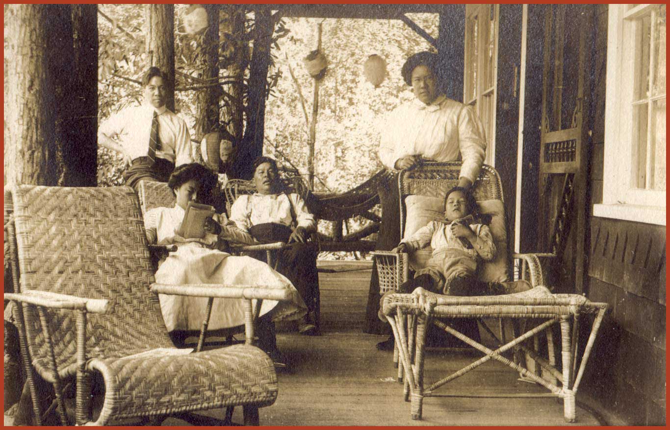 Black and white photo of a family sitting on the porch of their summer home. There is a young boy laying on a lawnchair and a young girl reading a book.