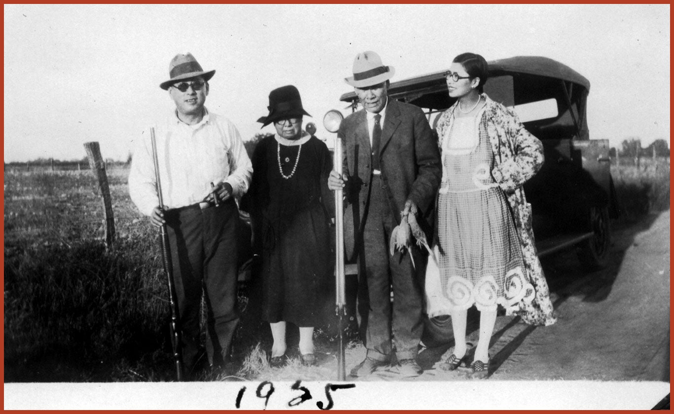 Joseph and Mary Tape stand side-by-side with their son Frank Tape, and his wife, Ruby. Joseph and Frank hold hunting rifles. Behind them is a car and a wife open field.
