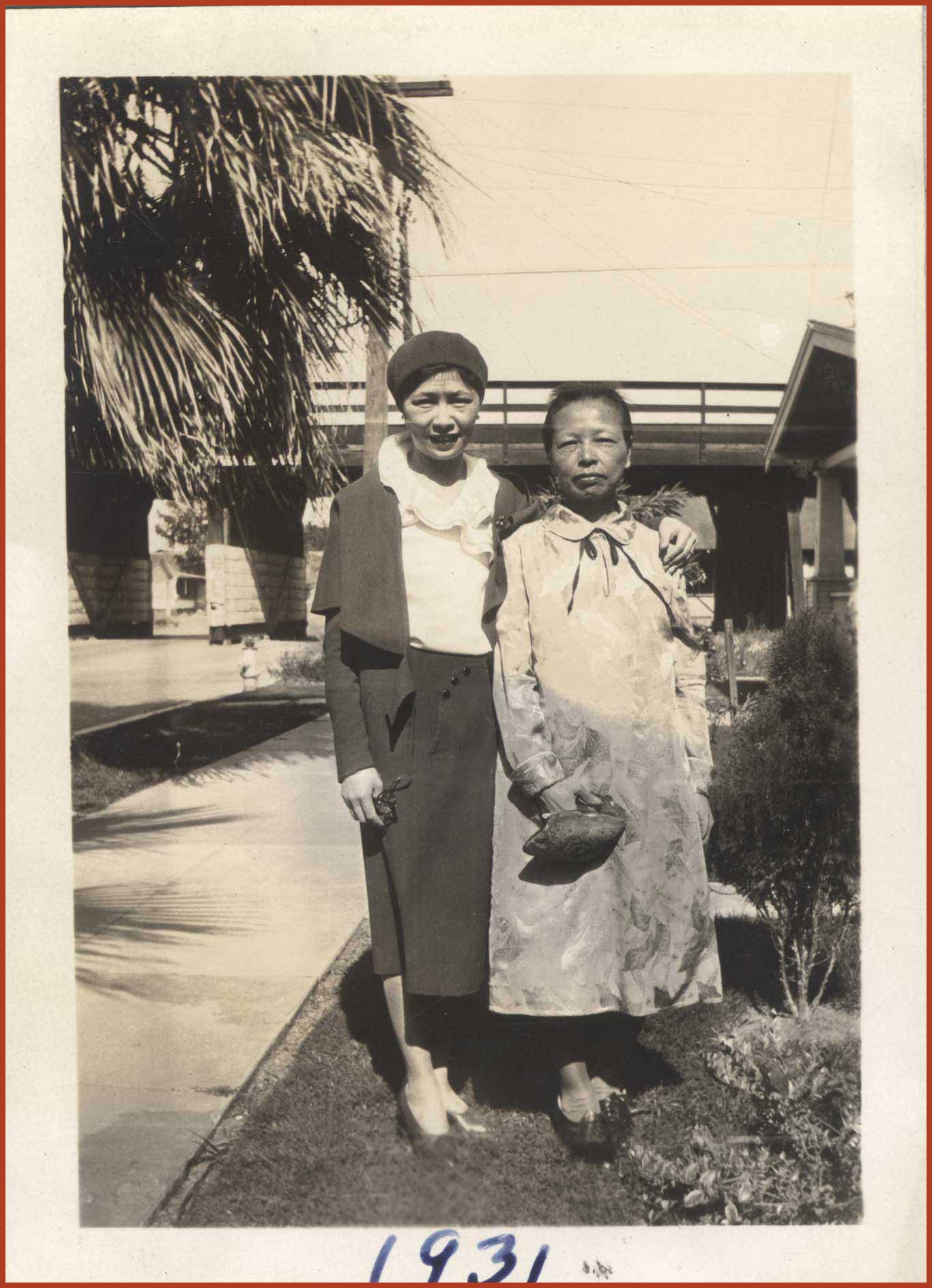 Black and white photo of a woman with her arm around her mother. They are standing in front of a building next to a palm tree.