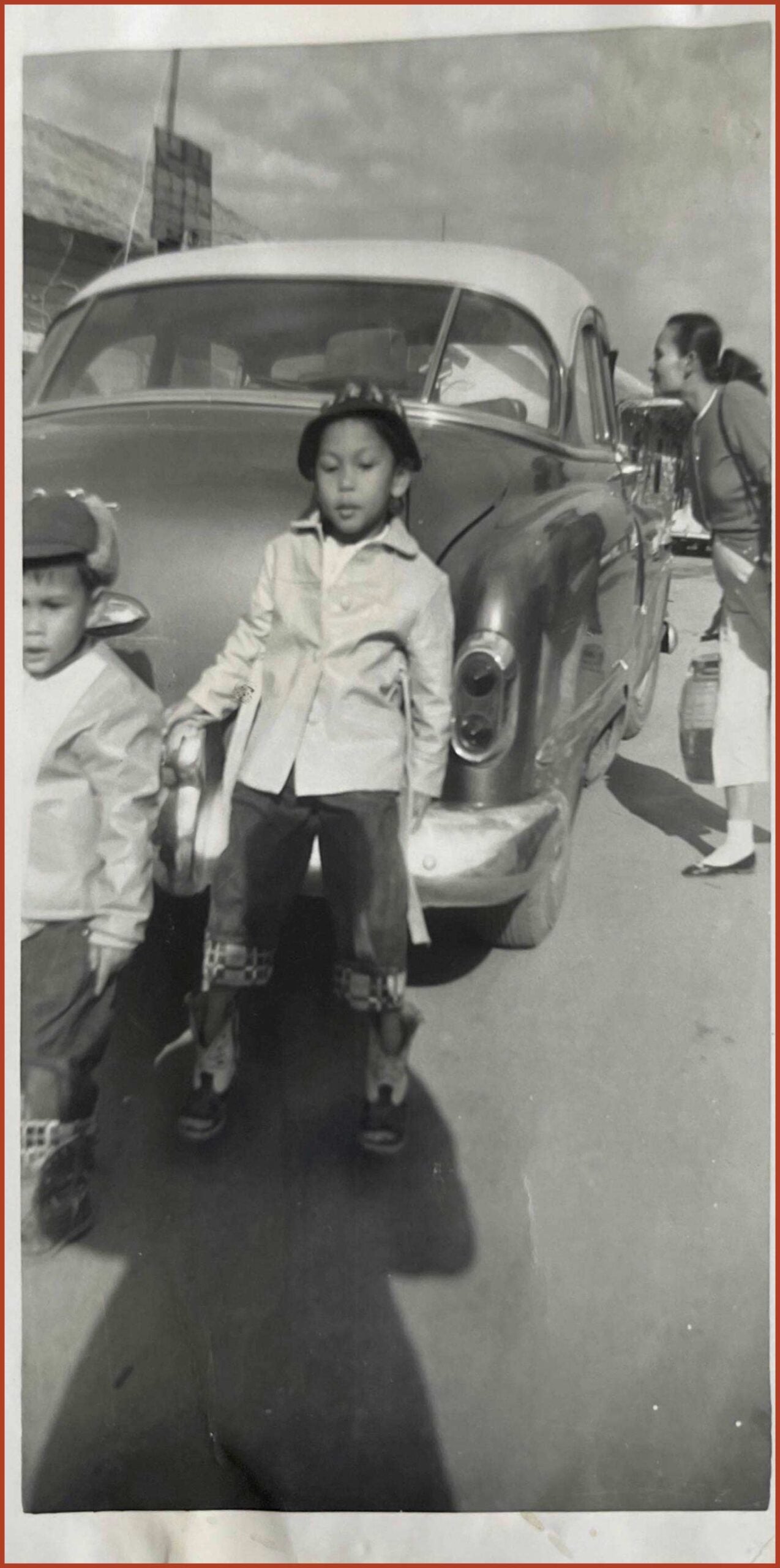 Two children standing in front of a car. A woman is standing next to the car speaking to someone inside of the passenger seat.