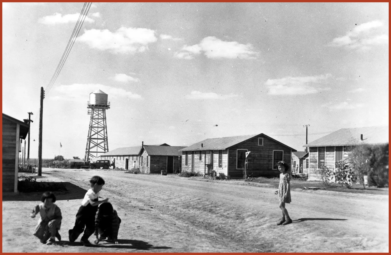 Three children playing outside. In the background are a few sparse buildings and a water tower.