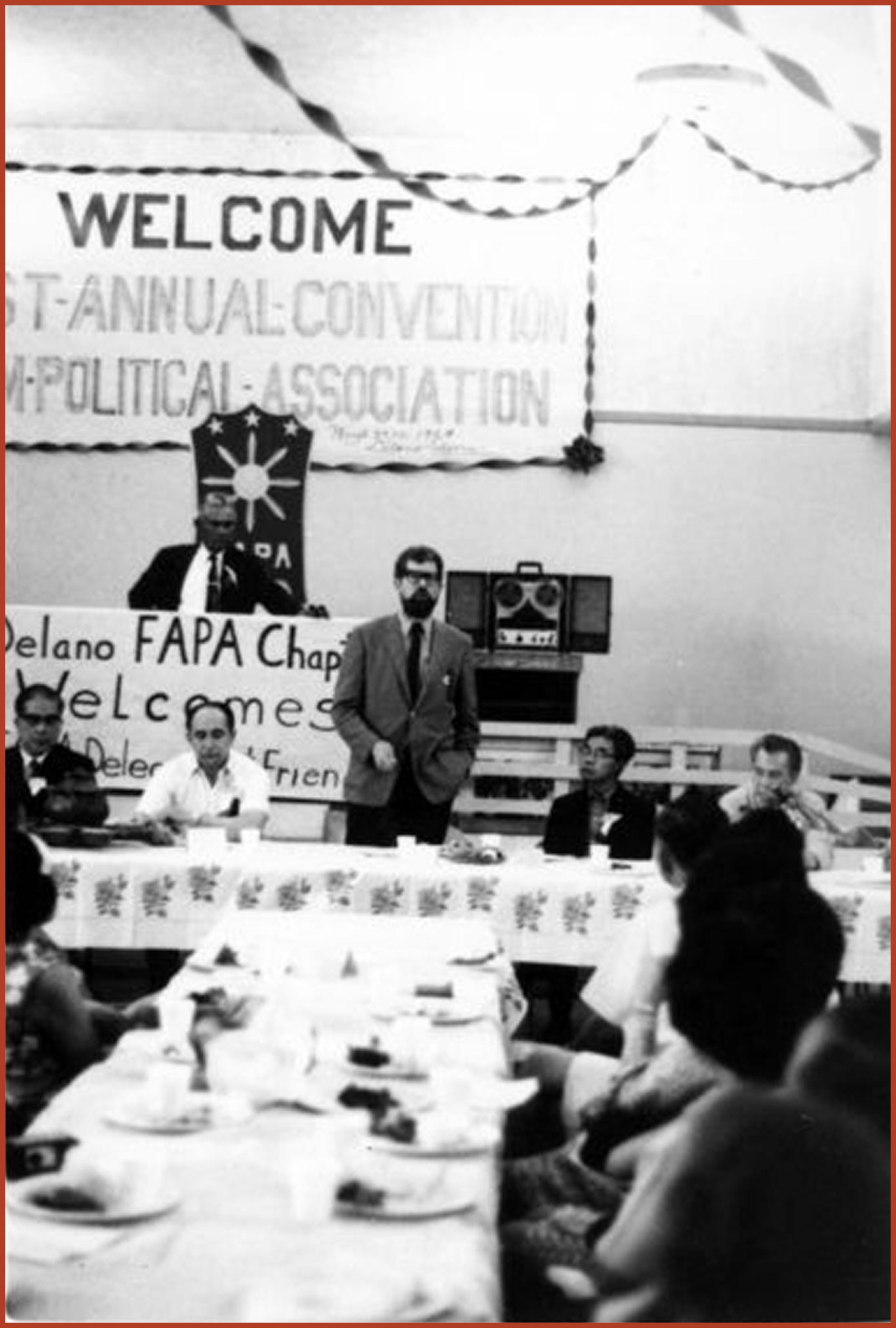 Group of individuals dining at a large table. A man is standing and speaking to the attendees. There is a banner in the back that says "Welcome 1st-Annual-Convention Filipino American Political Association."