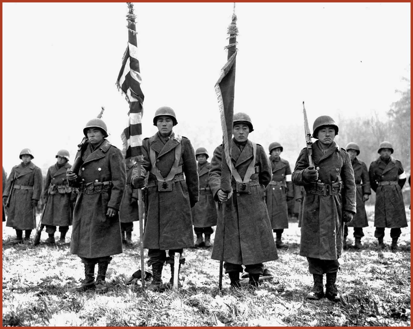 Two Color guards and color bearers of the Japanese-American 442nd Combat Team, stand at attention while their citations are read. Behind them, the rest of their combat team stand at attention.