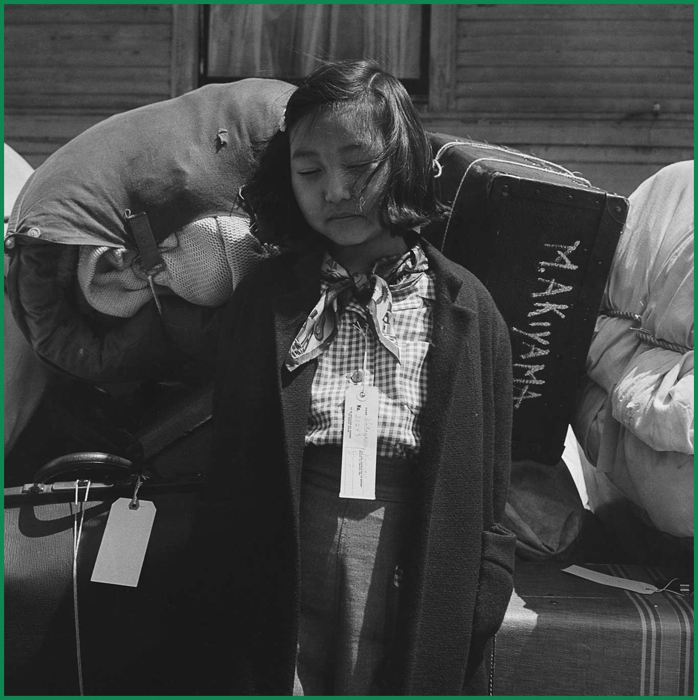 Young Kimiko Kitagaki waits in Oakland for a bus to transport her to a government camp, with tags with her name and government-assigned family number attached to her suitcase.