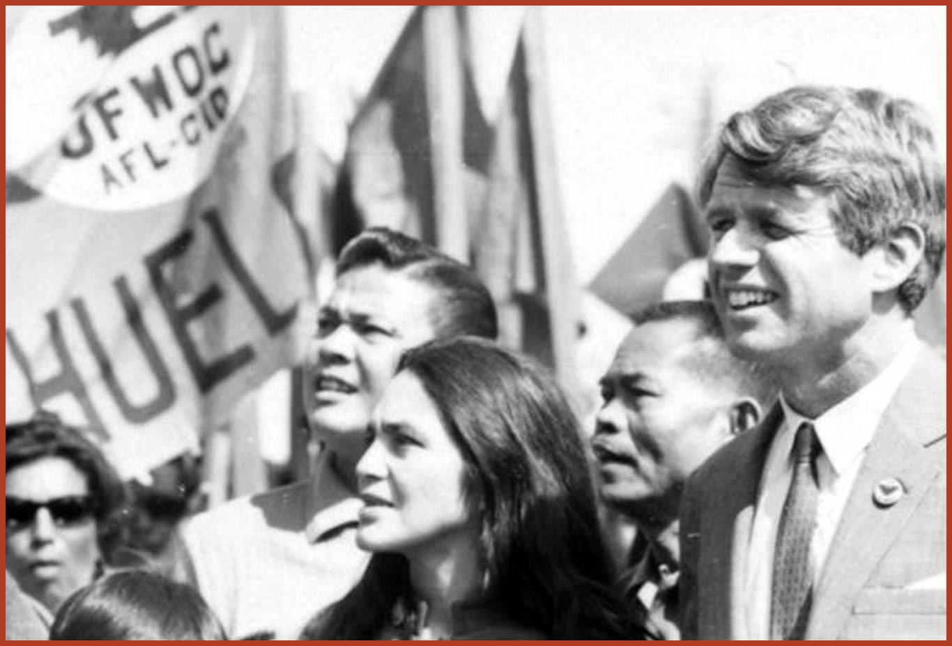 Black and white photo of 4 individuals looking off to the left. The United farm Workers Organizing Committee flag waves in the background.