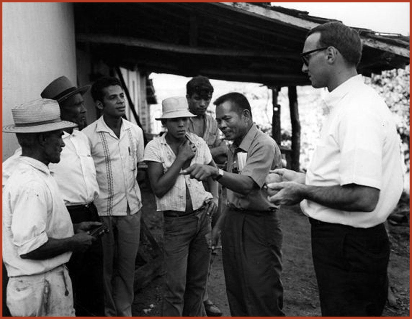 Larry Itliong and Dick Ginnold and a group of Brazilian farm workers talk together in front of a building.