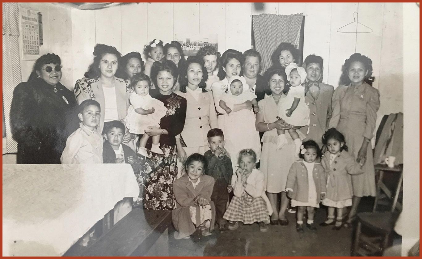 A group of women and their children pose for a photo in their bunkhouse. The group is dressed for a baptism.