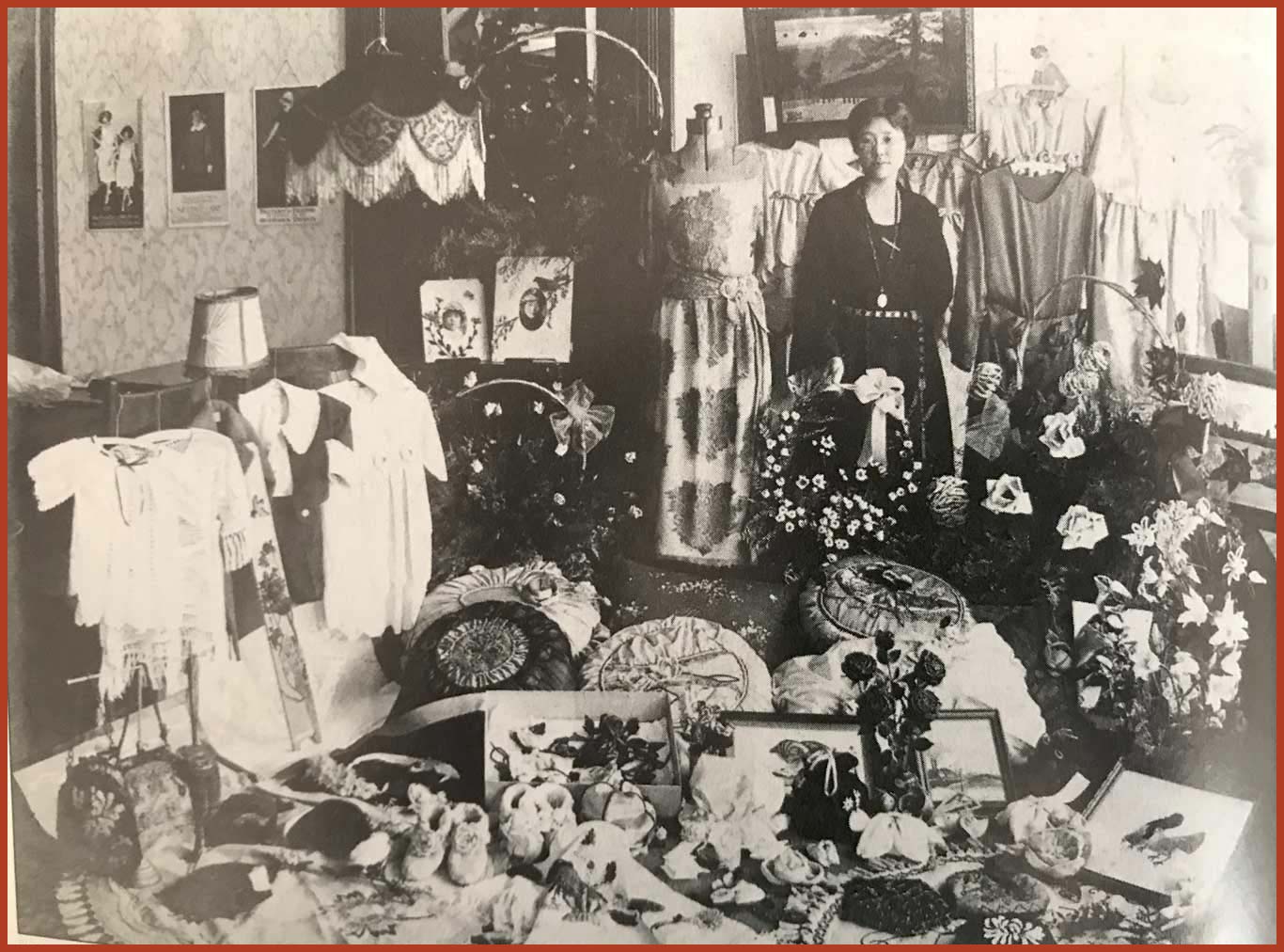 Black and white photo of a woman standing amongdt many dresses, flowers, and sewing equipment.