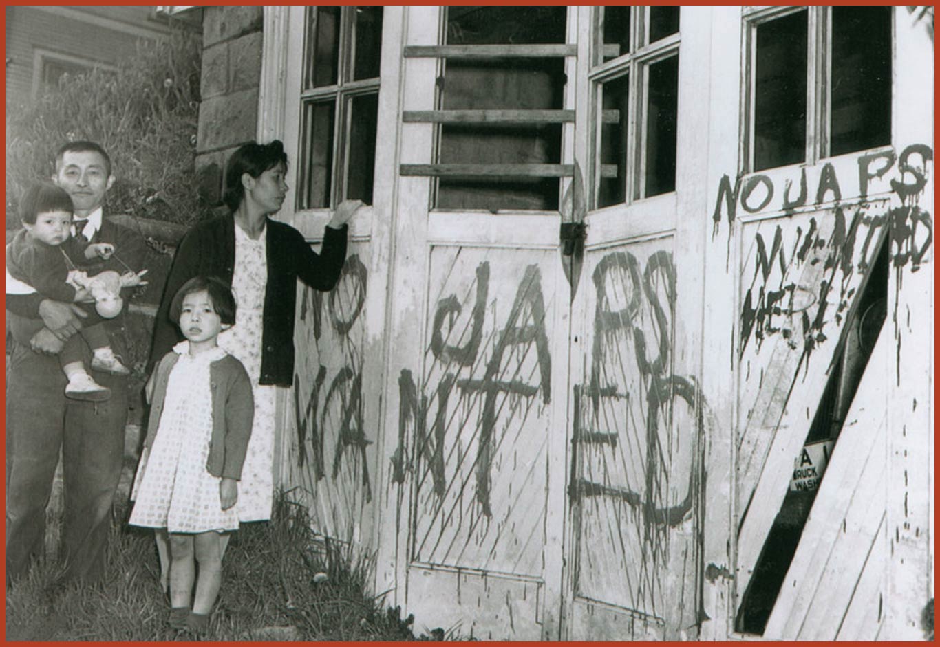 Nagaishi family standing in front of their garage that is vandalized with the words "No Japs Wanted." The father is holding a young boy in his arms while standing next to his wife and young daughter.