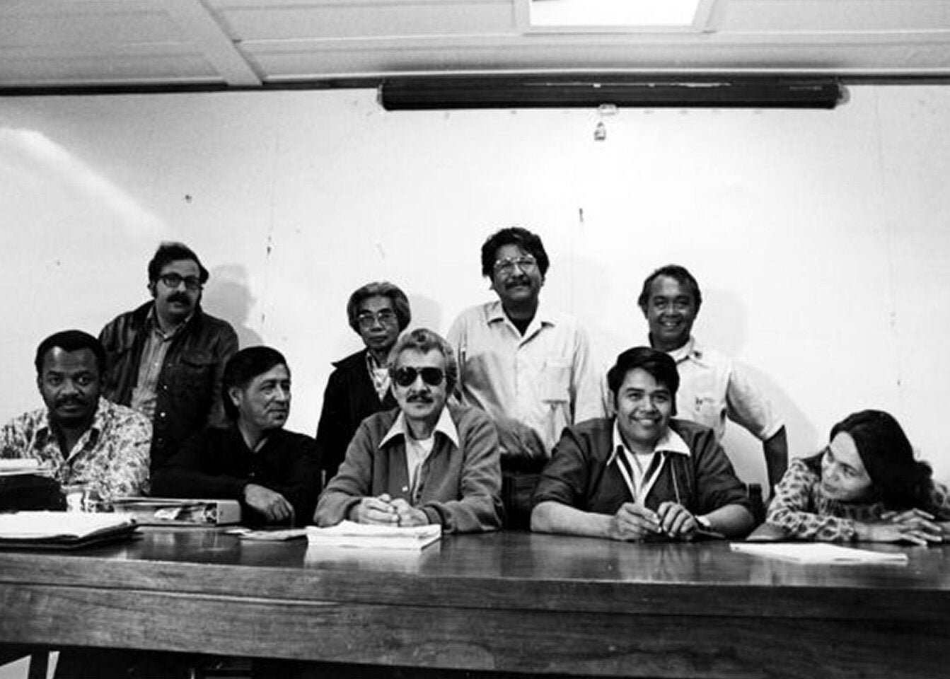Group of individuals gathered around a table. A man and a woman are speaking to each other from across the table.