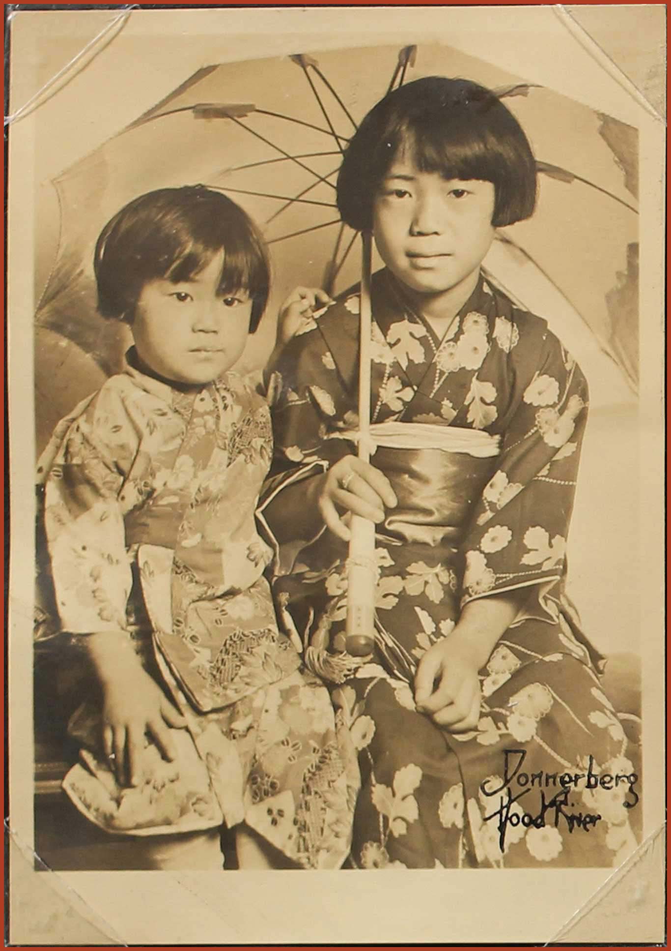 Yuka and Michi Yasui sitting, and wearing kimono. Michi is holding a cloth parasol.