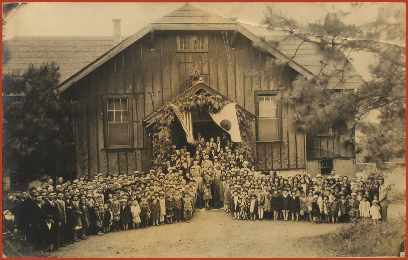A large group of Nikkei people stand in front of a large building. Coming out the front of the building, is the American flag on the left na dthe Japanese flag on the right.