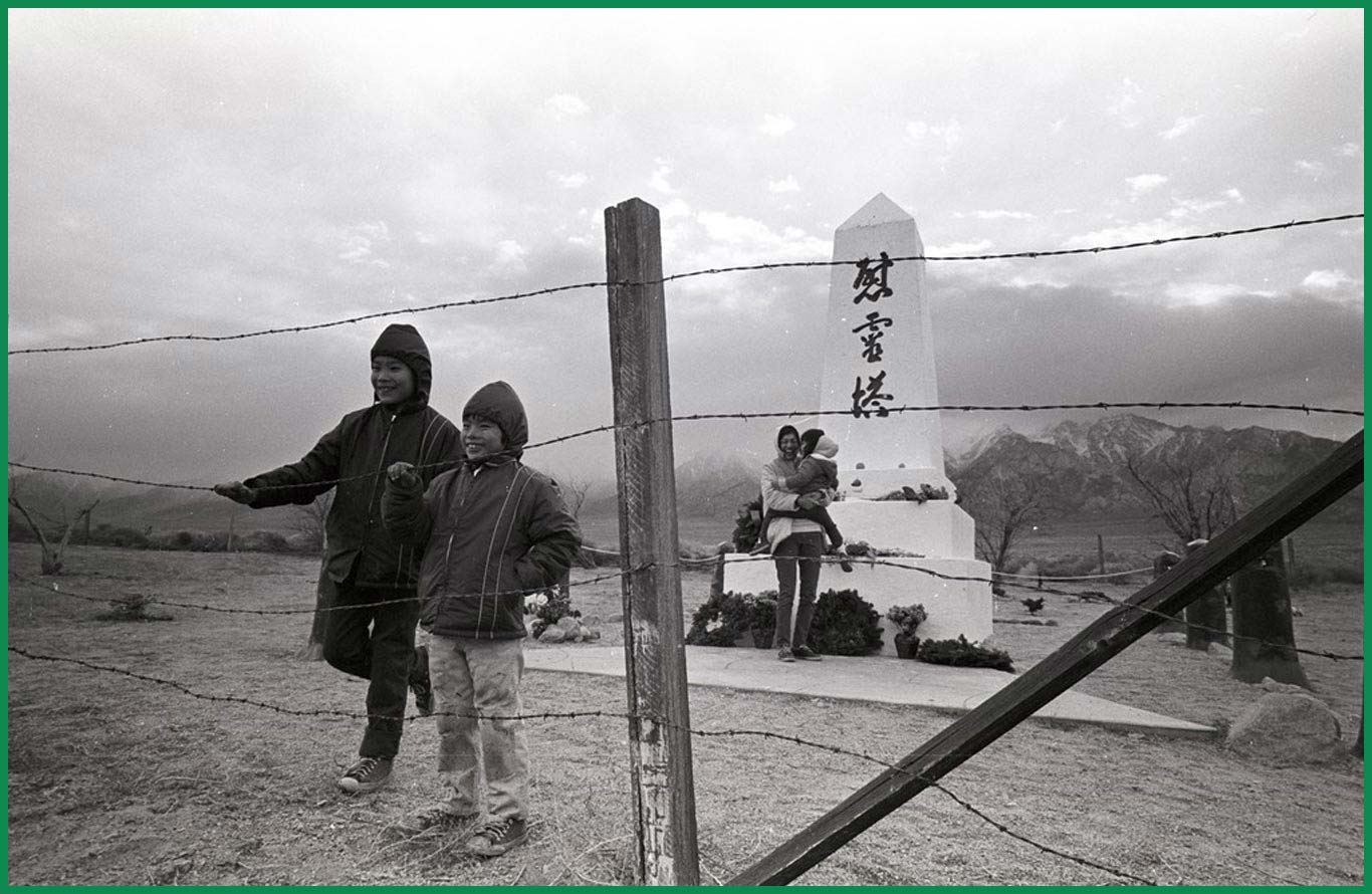 Two children are standing and holding onto a wired fence. Their mother stands in the back holding a baby. In the back is the Manzanar Cemetery Monument, an obelisk painted with Japanese characters.