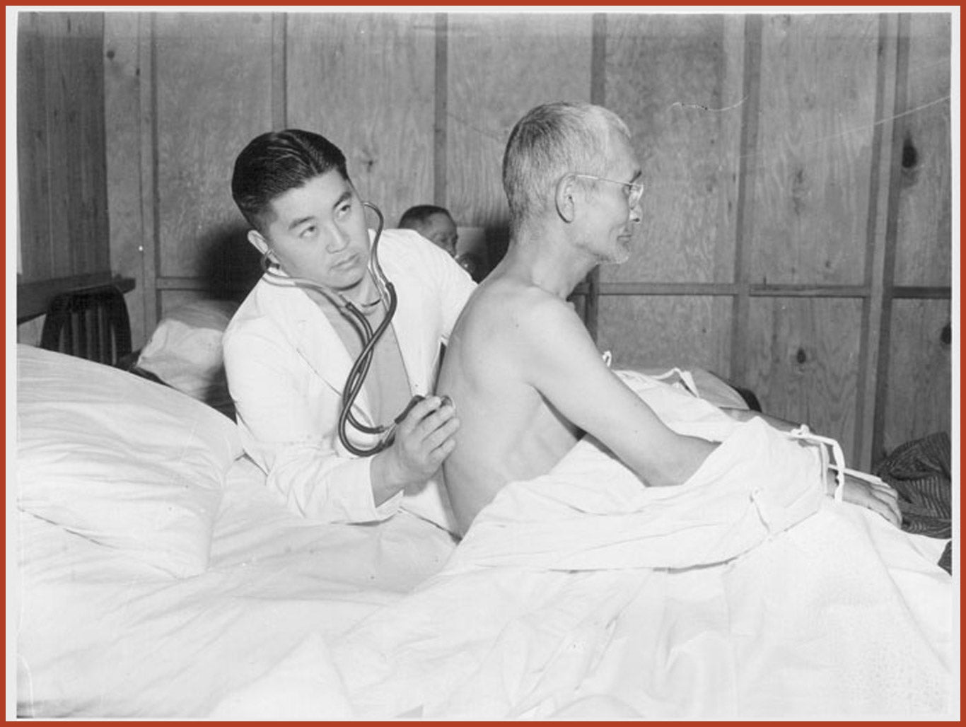 Doctor in white coat examines an elderly man in a bed by using a stethoscope on his back.