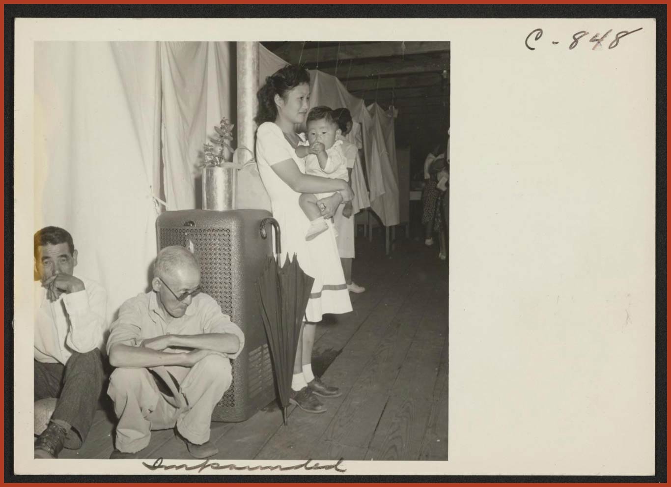 A group of people huddled together at the Manzanar Relocation Center. Behind them a line of short white cloths offer a small amount of privacy.