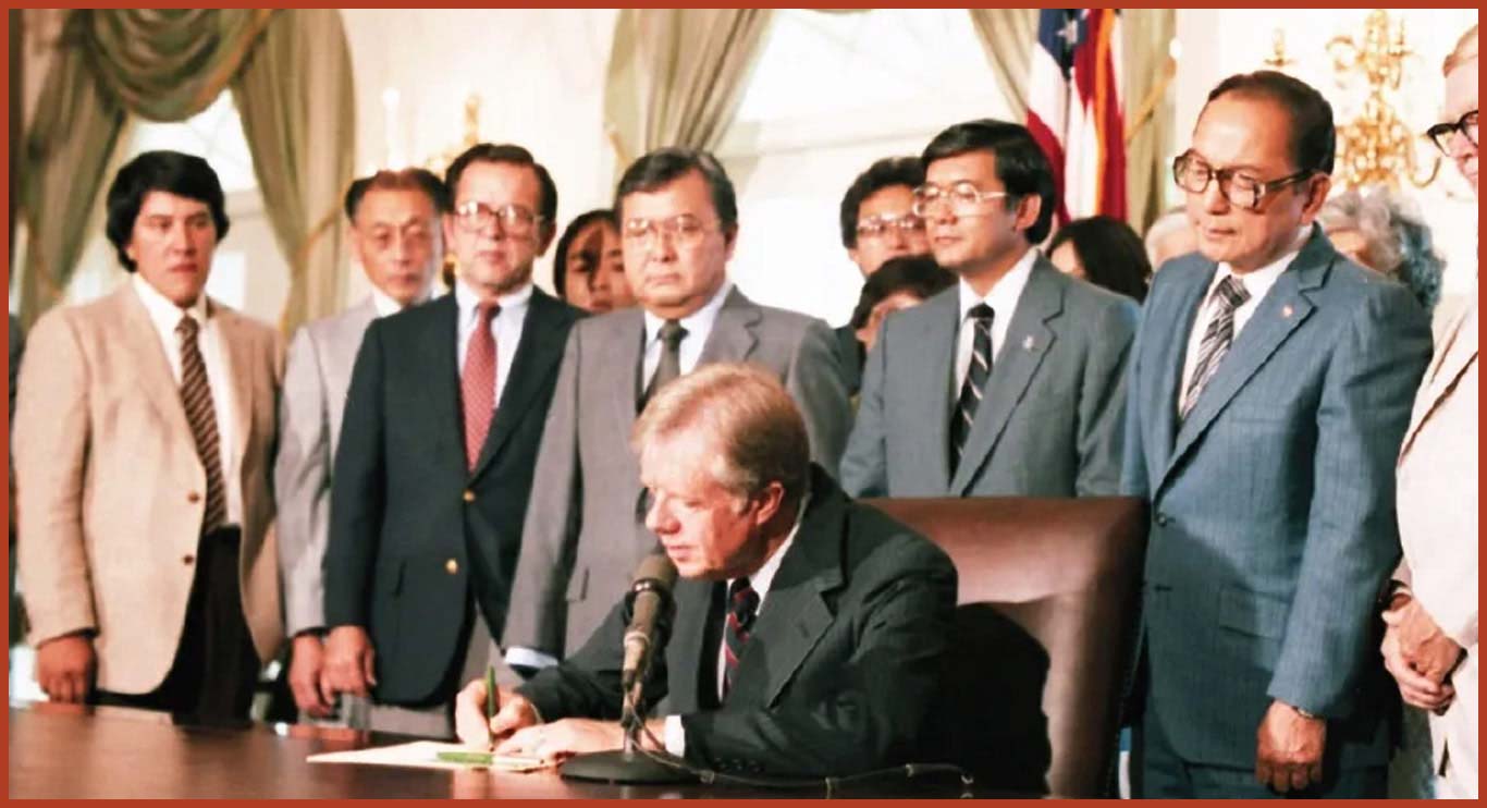 Clifford Uyeda, Ted Stevens, Daniel Inouye, Ron Ikejiri, Barbara Ikejiri, Norm Mineta, Spark Matsunaga, George Danielson, stand behind a seated President Carter as he signs the Commission of Wartime Relocation and Internment of Citizens Act.