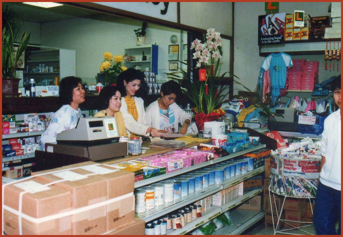 Four women behind a cashier counter in a pharmaceutical store. A man stands off to the side in front of the counter. The women are smiling and talking.