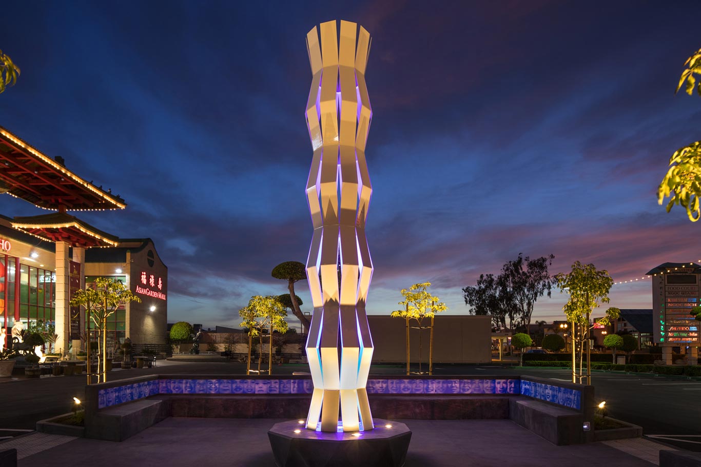 A tall, geometric light sculpture with a twisting, segmented design glows with soft white and purple lighting at dusk. The sculpture stands at the center of a low, angular fountain with blue tile accents. In the background, there are small trimmed trees, a building with a traditional Asian-style roof, and signage reading "Asian Garden Mall," all under a dramatic evening sky with blue and purple clouds.