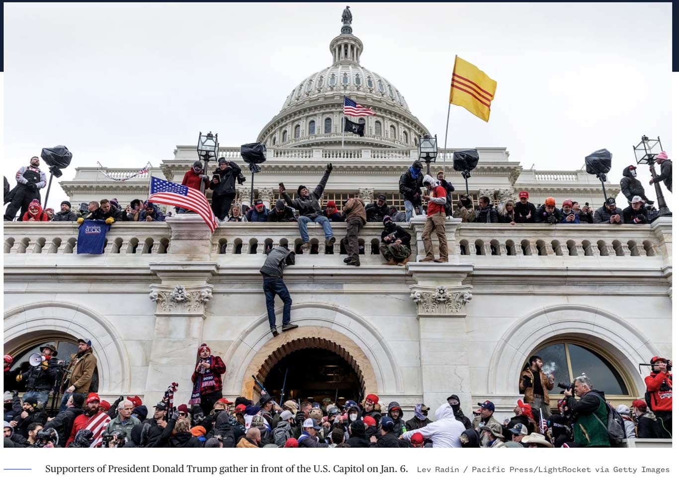 Many individuals climbing the walls of the Capitol Building in Washington D.C. while holding the American flag, microphones, rifles, and cameras. Many adorn hats that read "Make America Great Again."