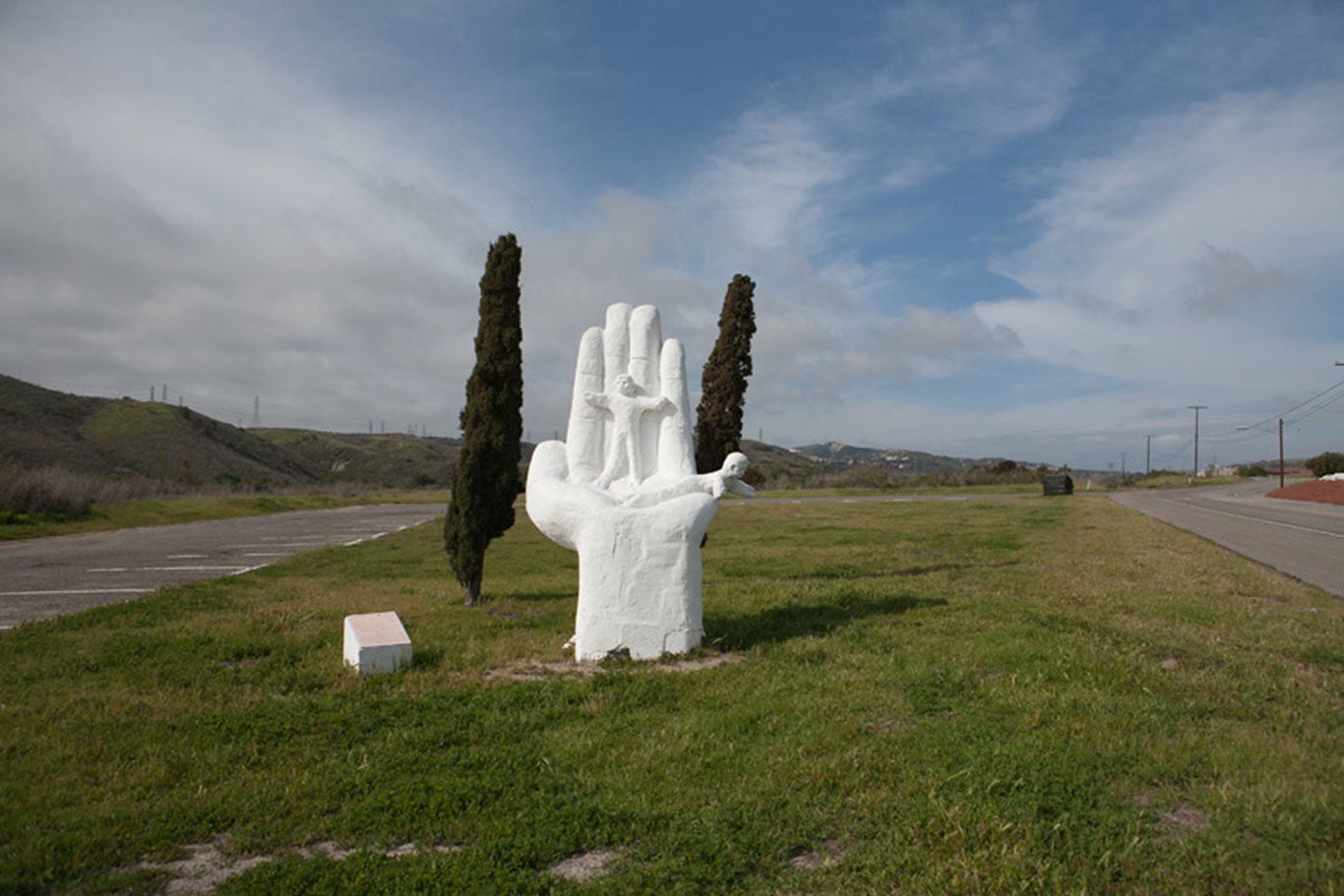 Large white cement statue of an open palm with a little girl standing along the fingers while a little boy lays down on the palm.