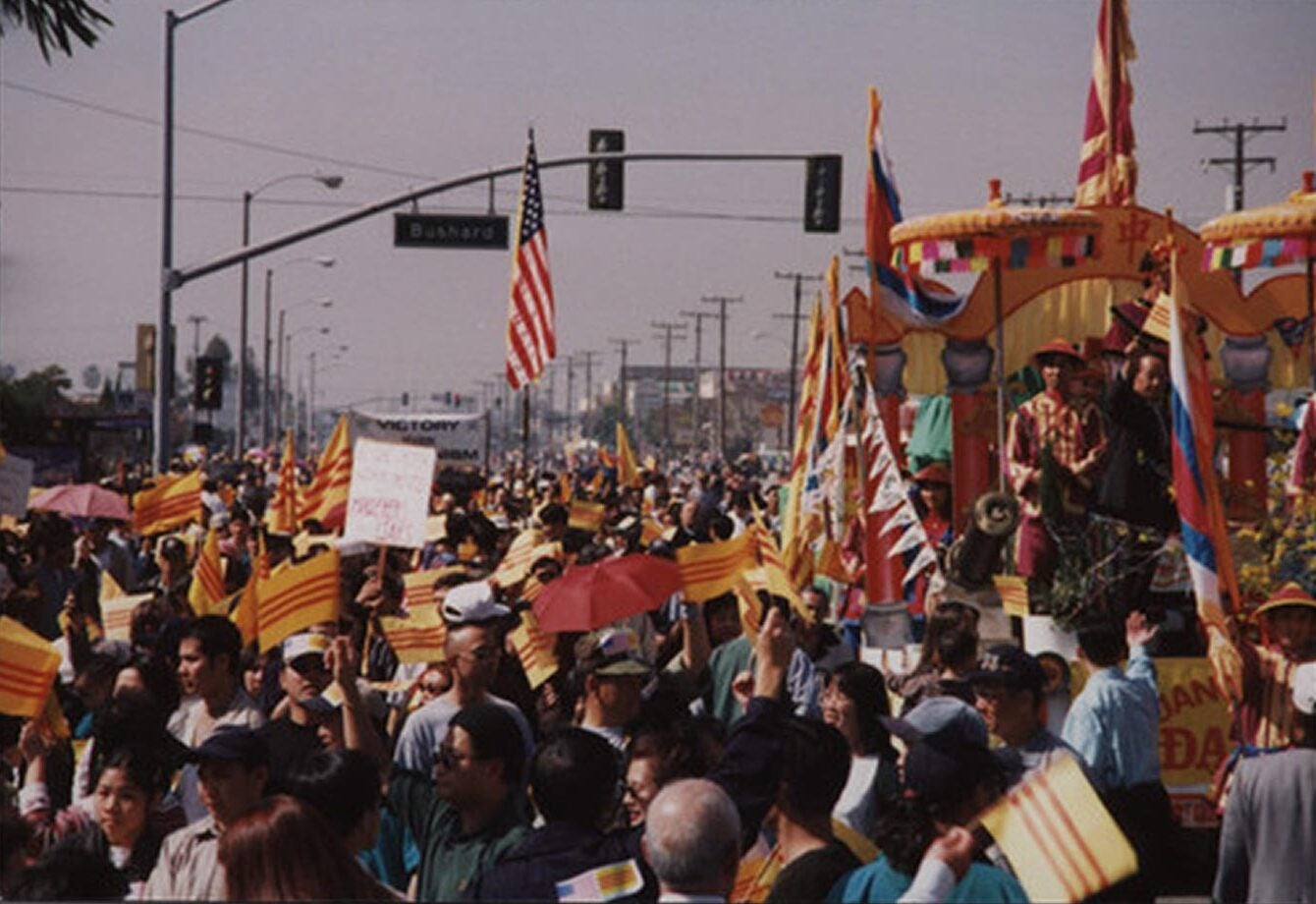 Many individuals parading in the street waving the American flag, South Vietnamese flags, and other signs. There is a float decorated in yellow colors and participants on the float adorn traditional Vietnamese attire.