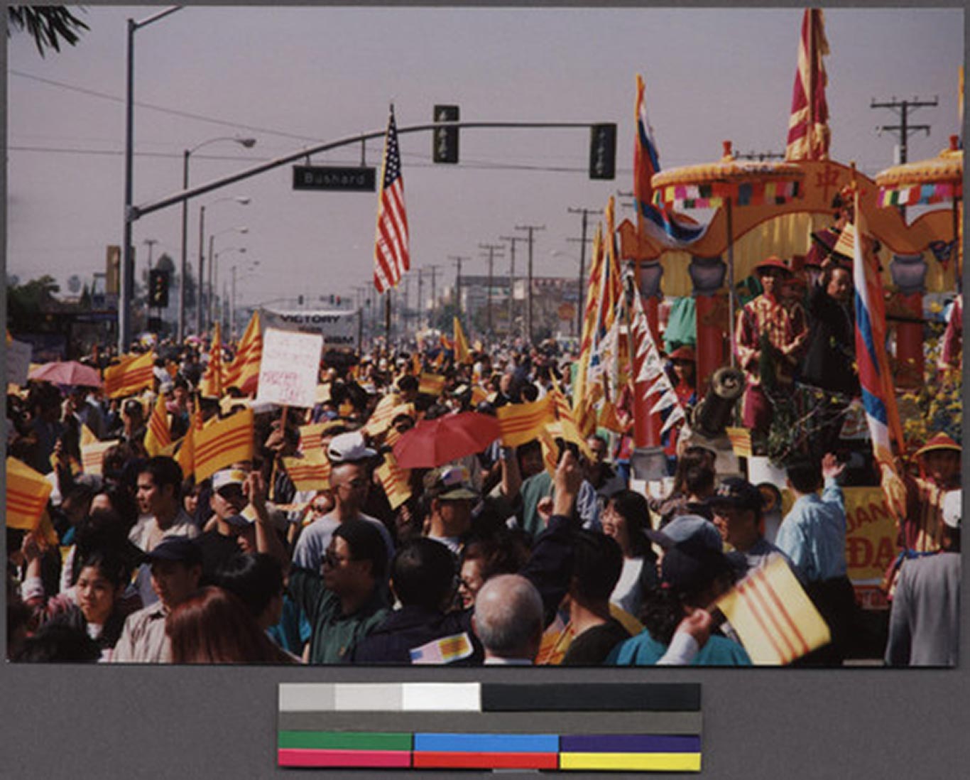 Many individuals parading in the street waving the American flag, South Vietnamese flags, and other signs. There is a float decorated in yellow colors and participants on the float adorn traditional Vietnamese attire.