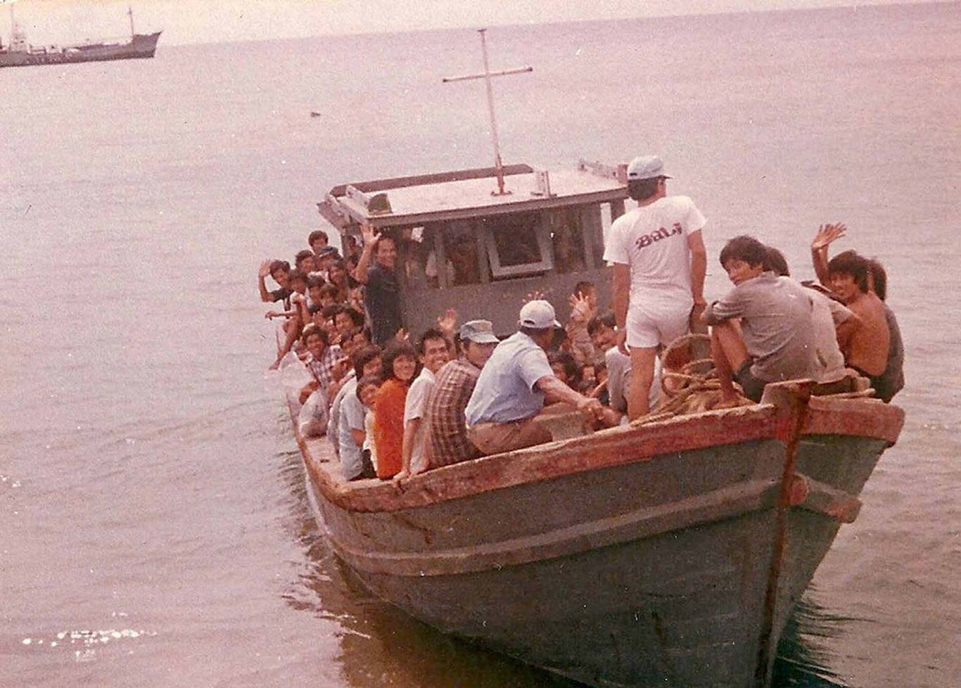 A color photograph of a crowded wooden boat on calm water, carrying many people sitting and standing along the edges and on the deck. The passengers, including adults and children, are wearing casual clothing and appear to be waving or looking toward the camera. The boat is close to shore, and in the background, another larger ship is visible on the horizon.