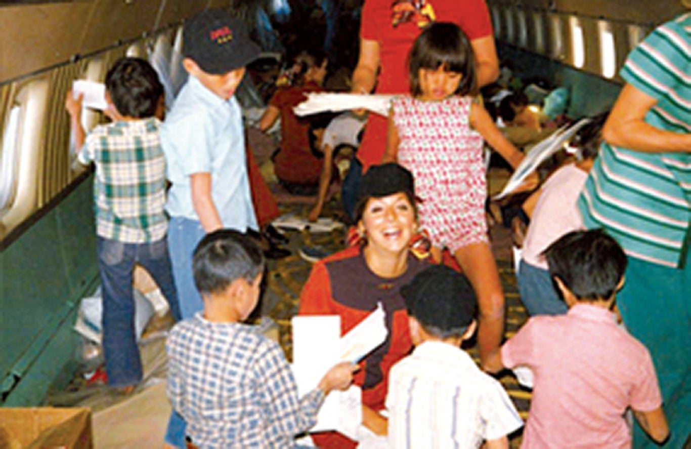 Many children play inside of an airplane as flight attendants sit amongst them and assist them. The flight attendants adorn red dresses.