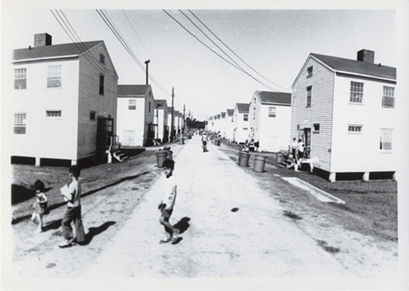 Black-and-white photo of a dirt road lined with houses. Three children crossing the road turn to face the camera.