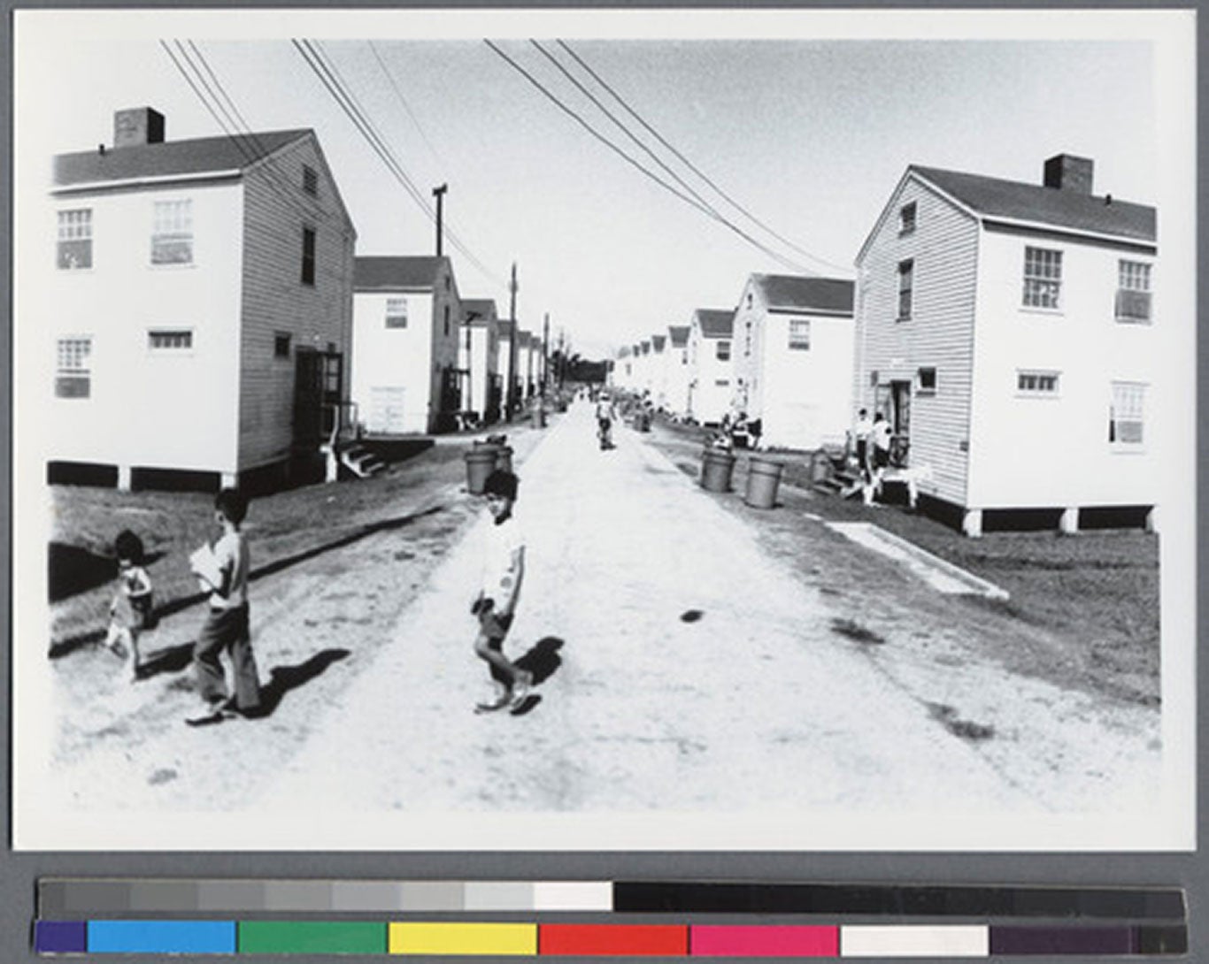 Black-and-white photo of a dirt road lined with houses. Three children crossing the road turn to face the camera.