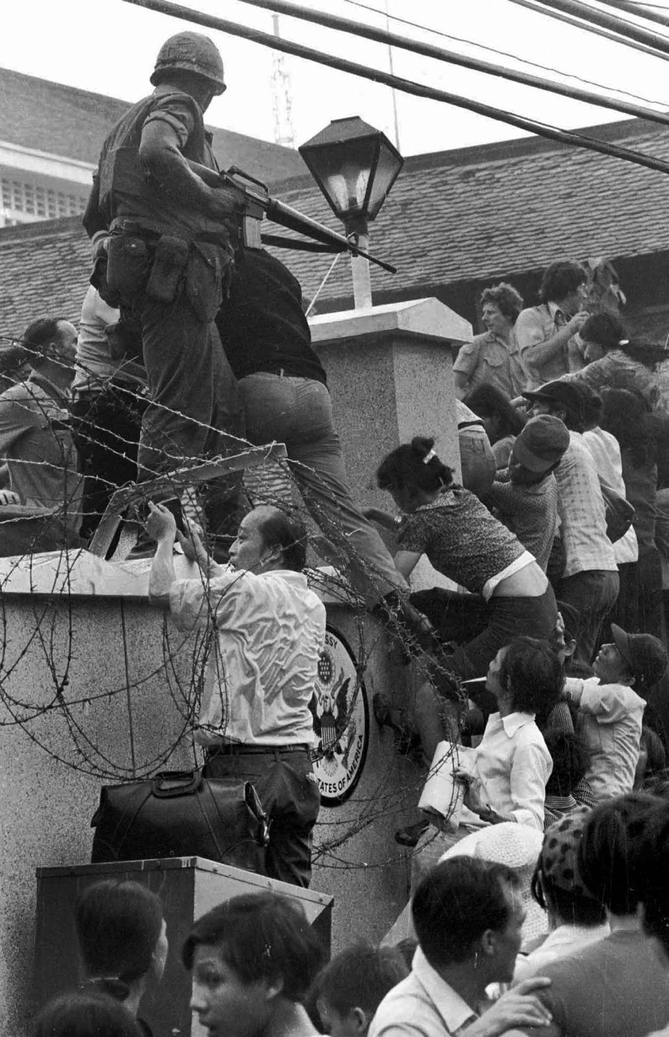 Black-and-white photo showing many individuals climbing a wall and breaking a barbed fence. A soldier with a rifle stands amongst them.