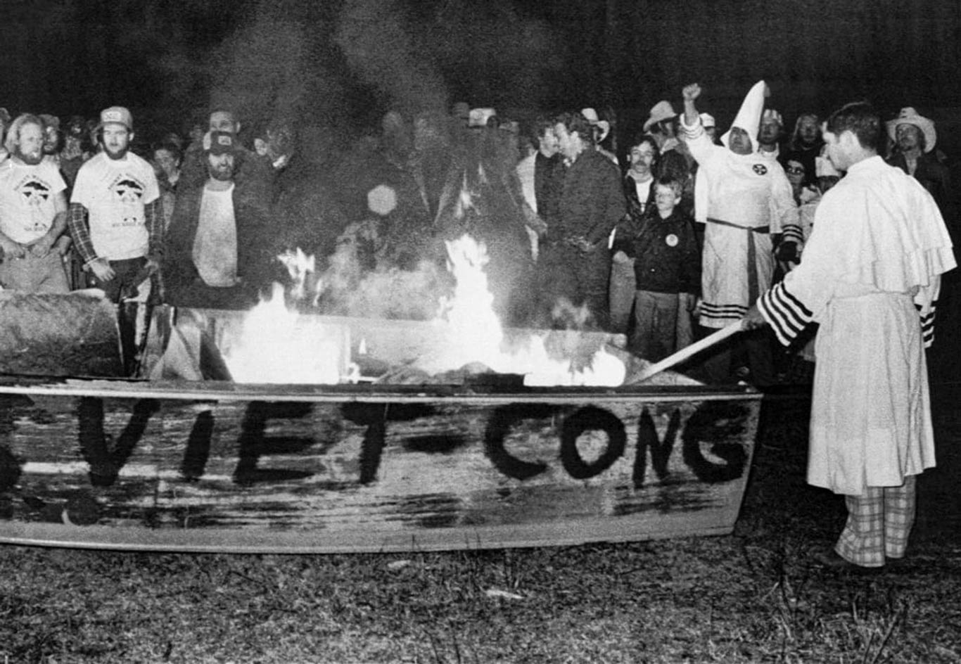 A large group of white men gather round as Ku Klux Klan members set fire to a wooden boat with the words "VIET - CONG" painted on the side.