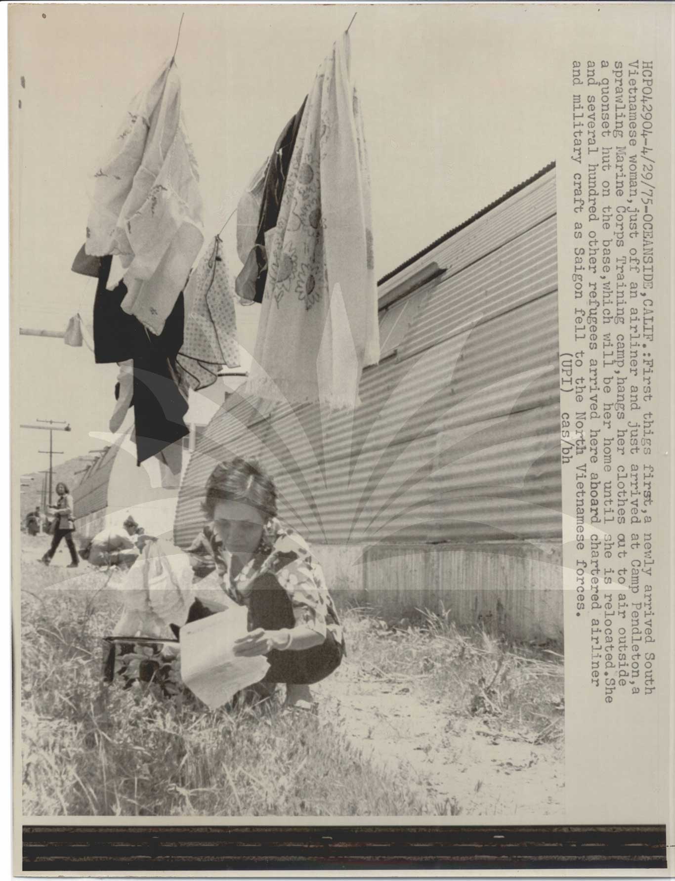 Black-and-white photo of a woman hunched over in the grass holding a bag and some papers. Above her, clothes dry on a clothesline.