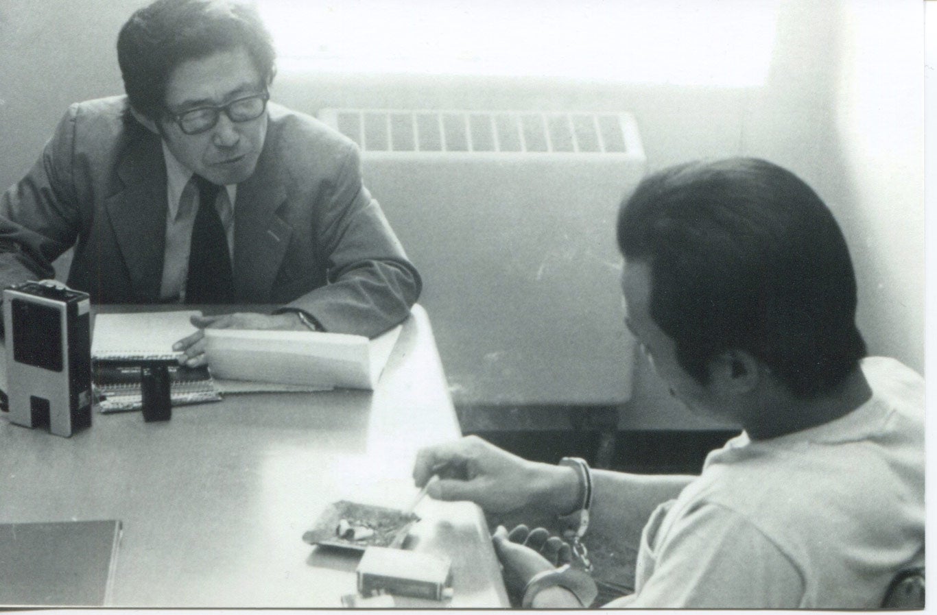 Chol Soo Lee handcuffed and smoking while speaking to K.W. Lee. Lee has notepads and other papers in front of him.