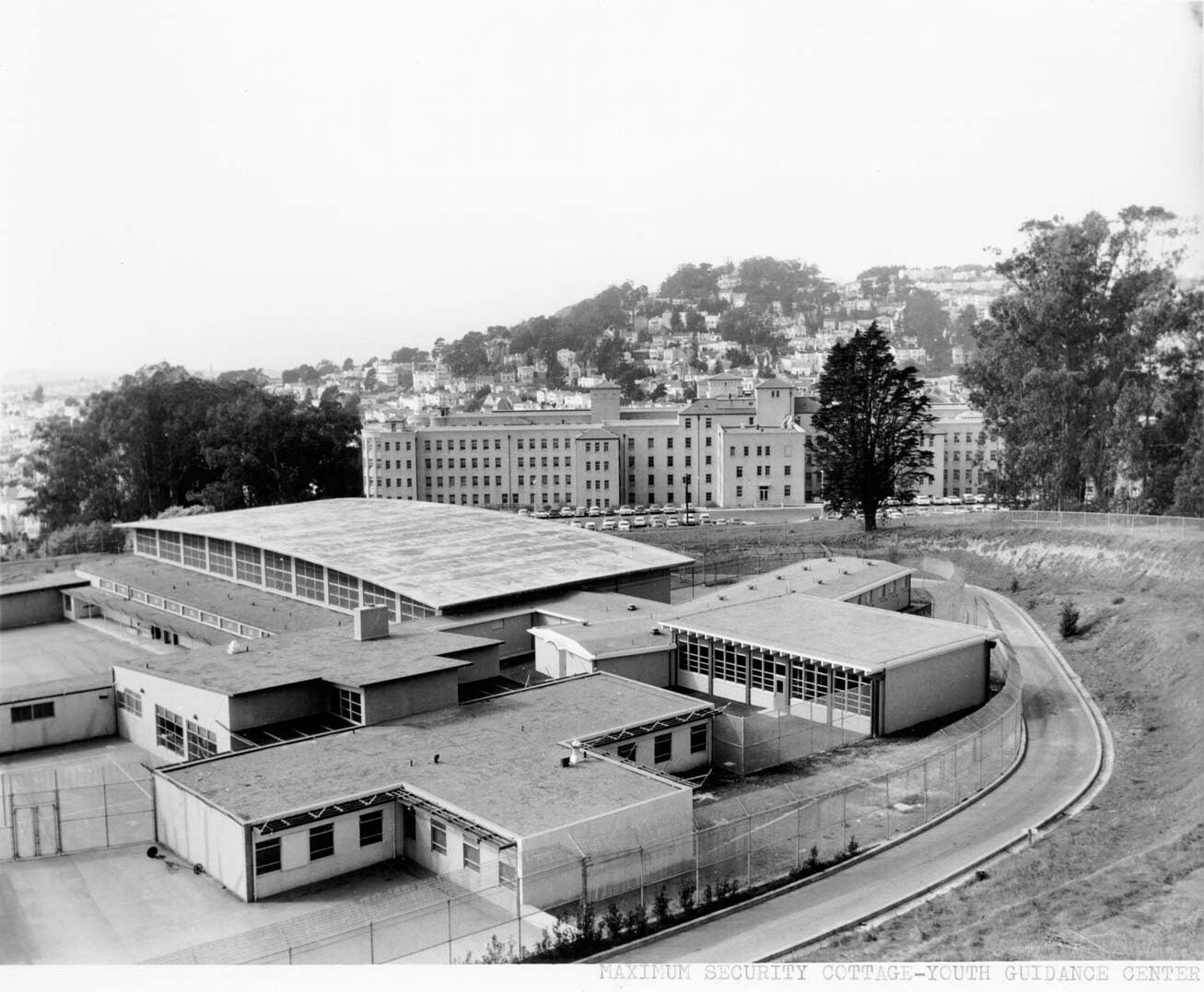 A black-and-white photograph shows a large institutional complex labeled as the "Maximum Security Cottage - Youth Guidance Center." In the foreground, several low, flat-roofed buildings are surrounded by a tall chain-link fence topped with barbed wire, indicating a secure facility. The buildings have numerous windows and are arranged in a grid-like pattern, with paved walkways and a curved driveway running alongside the perimeter fence. In the background, a larger multi-story building sits on a hillside, with rows of parked cars in front and residential homes scattered up the slope behind it. Tall trees flank the scene, and the overall setting appears to be on the edge of an urban area, with a mix of institutional and residential architecture visible in the distance. The photo is taken from an elevated vantage point, providing a wide view of the entire complex and its surroundings. The image is monochrome, emphasizing the stark and orderly nature of the facility.