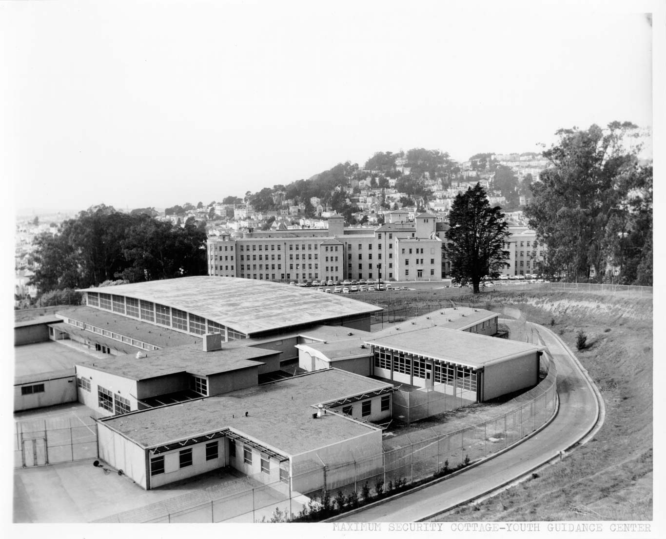 A black-and-white photograph shows a large institutional complex labeled as the "Maximum Security Cottage - Youth Guidance Center." In the foreground, several low, flat-roofed buildings are surrounded by a tall chain-link fence topped with barbed wire, indicating a secure facility. The buildings have numerous windows and are arranged in a grid-like pattern, with paved walkways and a curved driveway running alongside the perimeter fence. In the background, a larger multi-story building sits on a hillside, with rows of parked cars in front and residential homes scattered up the slope behind it. Tall trees flank the scene, and the overall setting appears to be on the edge of an urban area, with a mix of institutional and residential architecture visible in the distance. The photo is taken from an elevated vantage point, providing a wide view of the entire complex and its surroundings. The image is monochrome, emphasizing the stark and orderly nature of the facility.