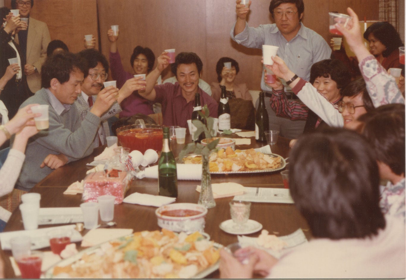 Chol Soo Lee sits at a table with a lot of food surrounded by many. All smile and raise a glass.