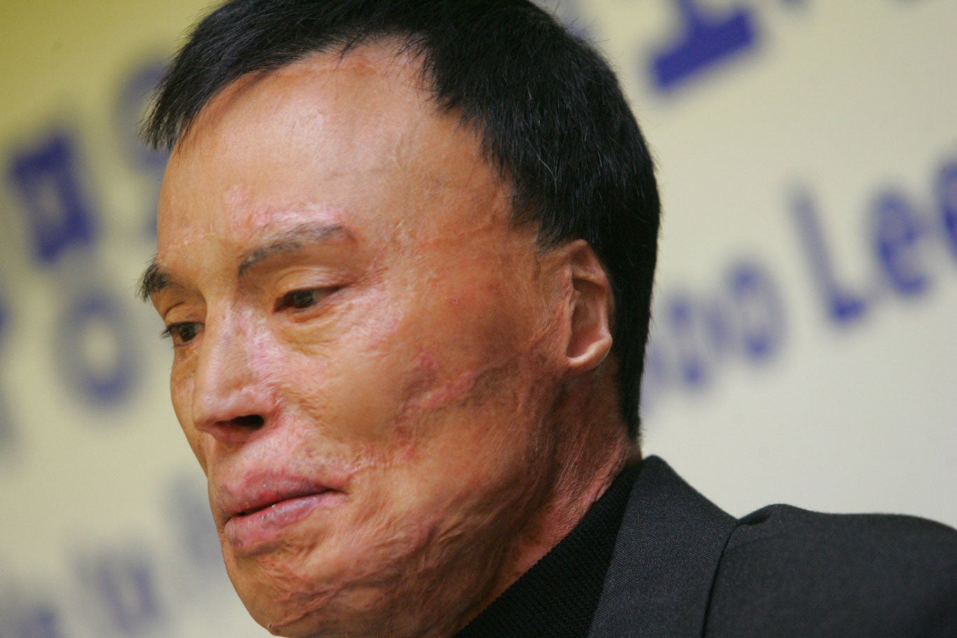 Chol Soo Lee with short, dark hair wearing a black jacket stands in front of a light-colored background featuring large, partially visible blue text. The image is closely cropped, focusing on this head and upper shoulders.