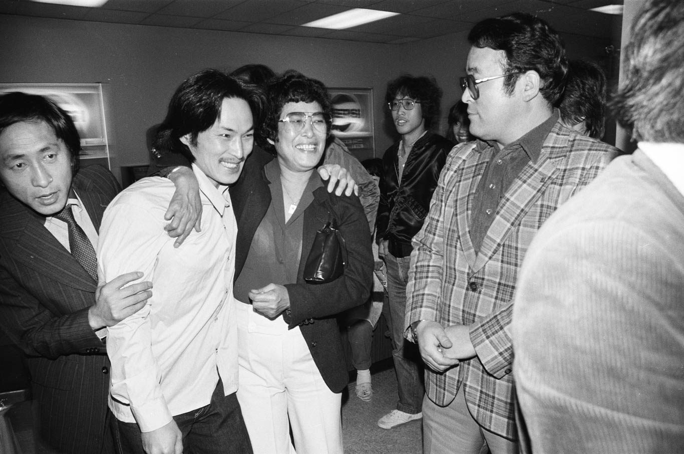 Chol See Lee hugging his friend with one arm. Other men are gathered around and chatting.