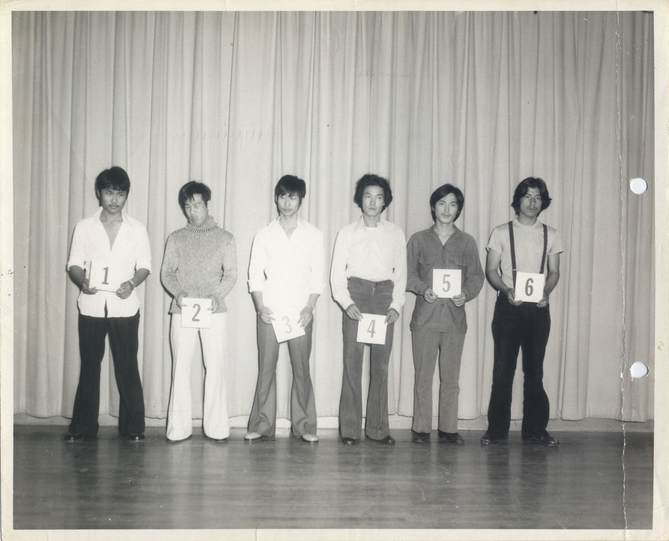Six men stand in a line on a stage in front of a curtain, each holding a card with a number, starting with 1 on the left and going through to 6 on the right. Suspect number 5 is Chol Soo Lee.